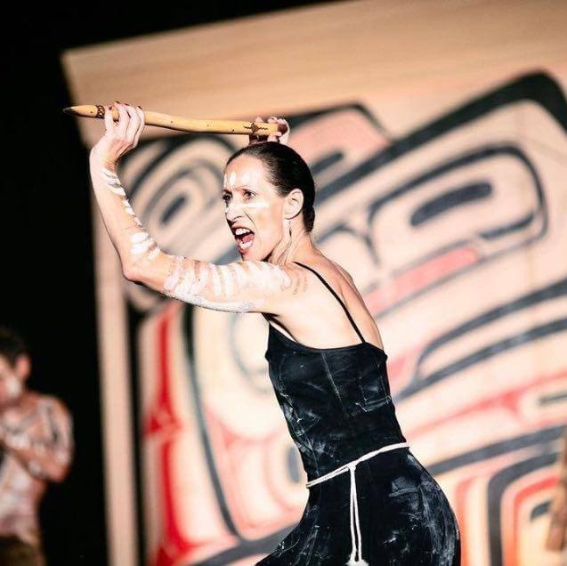 A woman holds a spear above her head with two hands. She is dressed in a black dress ad has chalk markings on her arms and face. 