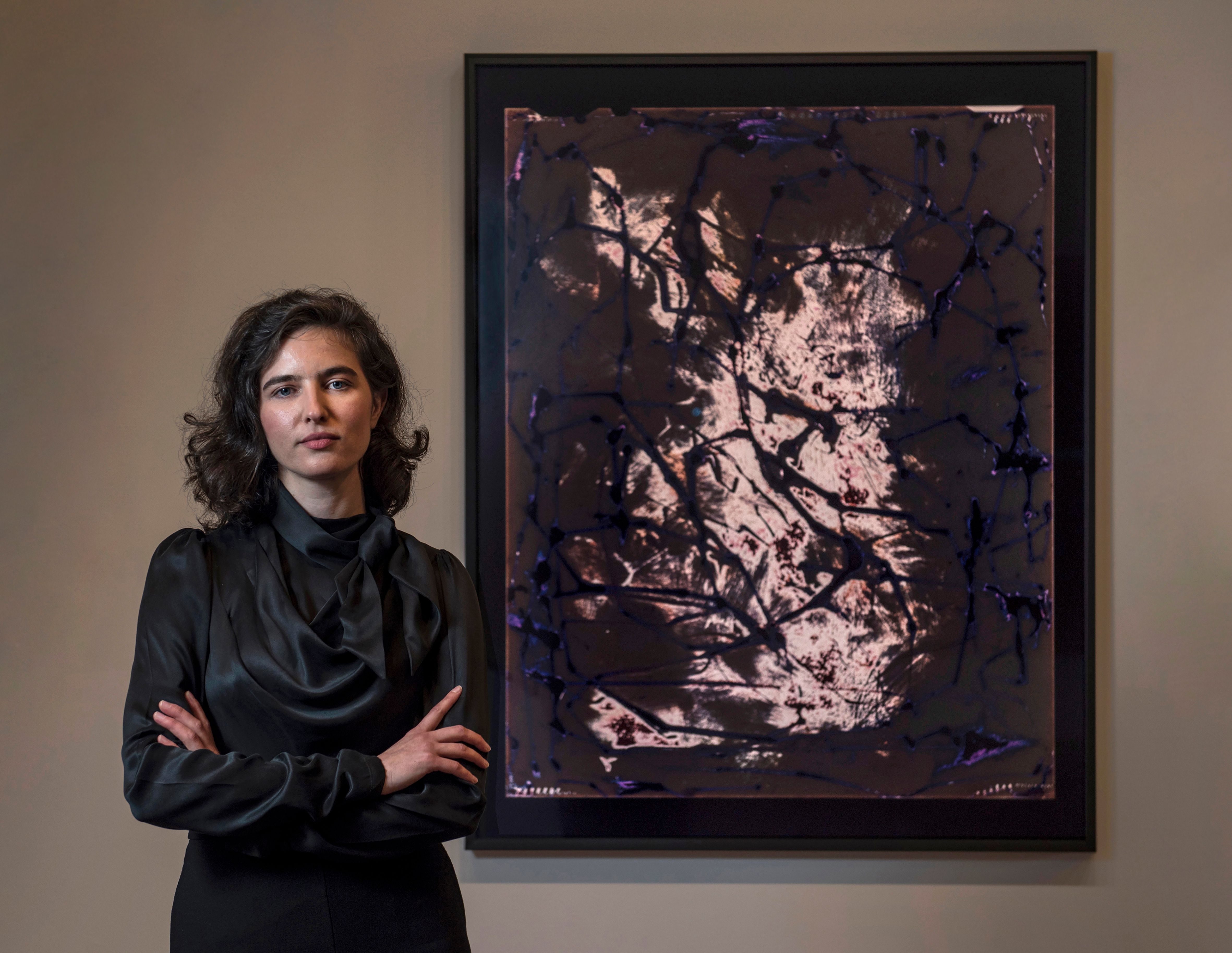 A woman wearing a black shirt looks at the camera with arms crossed. She is standing next to an abstract artwork.