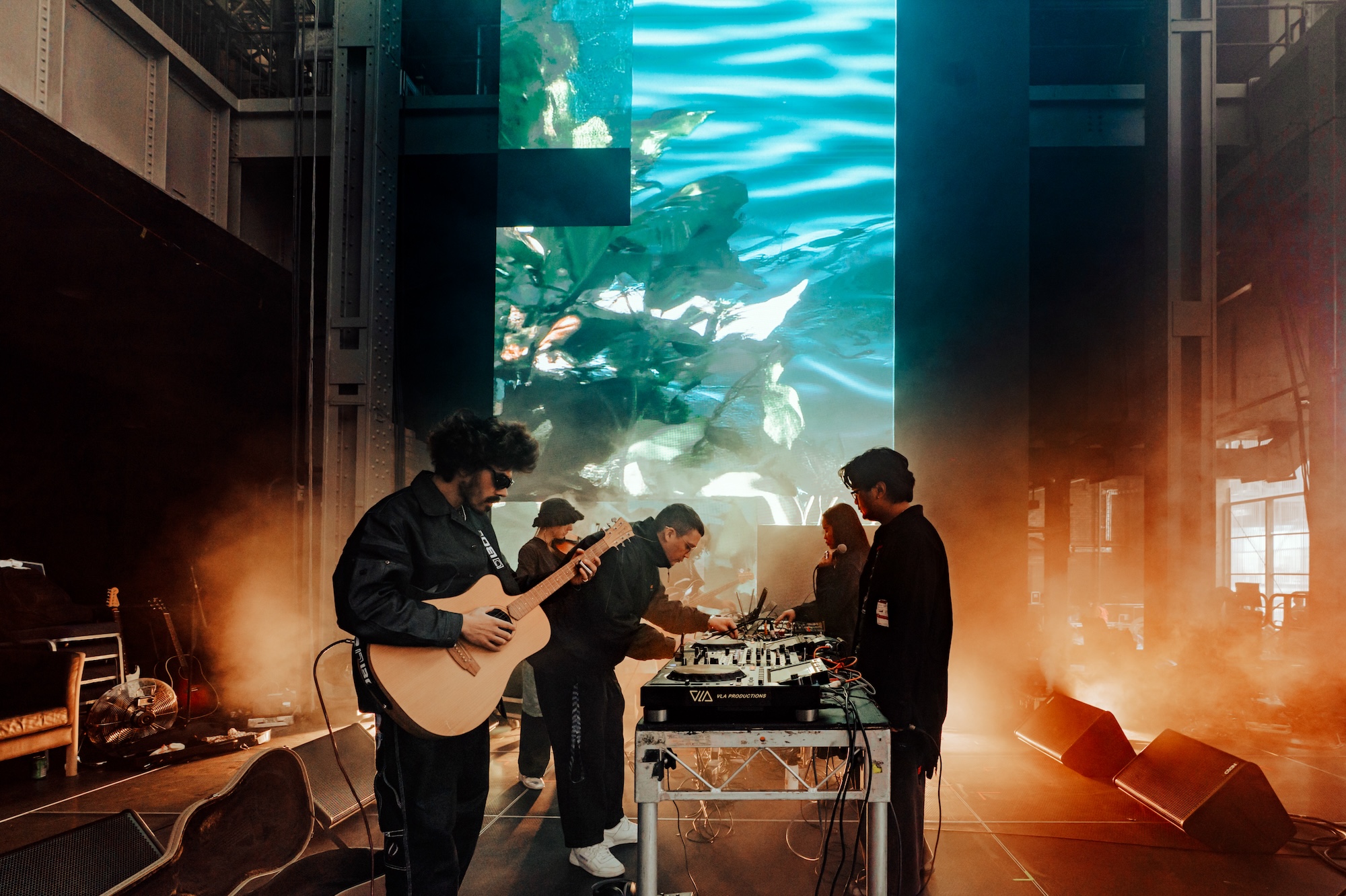 A photograph of an ensemble playing a guitar and mixing desk, with a screen behind them.