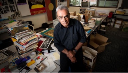 A photograph of Gary Foley sitting on a desk covered in books.