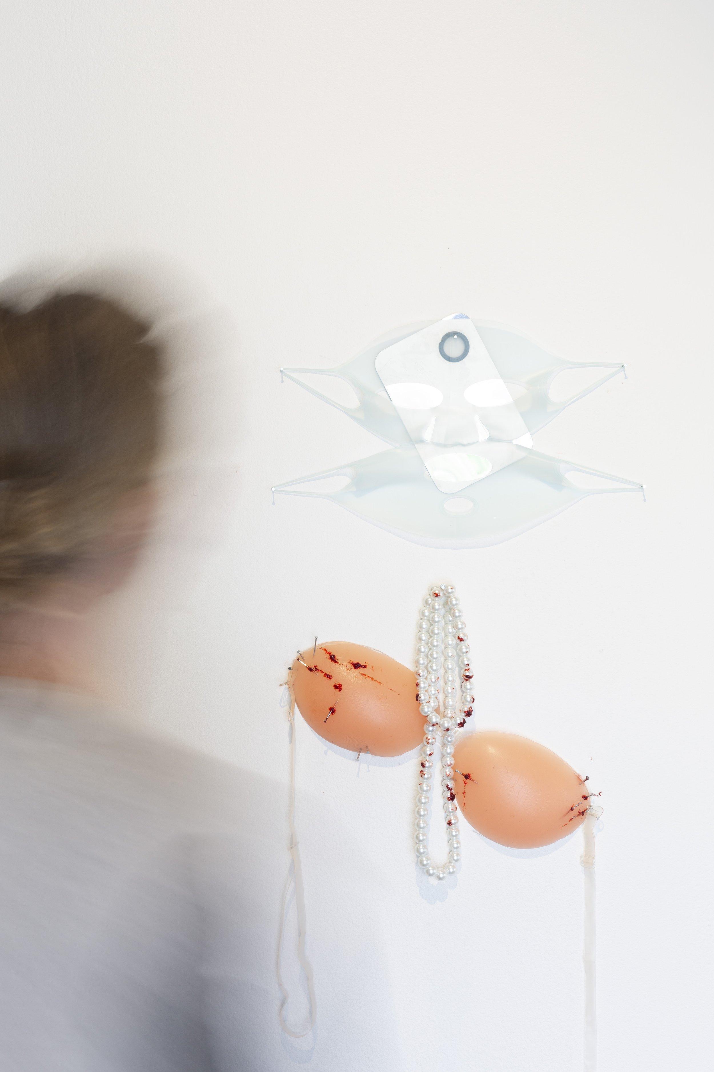 A person is blurred on the left. They are staring at a silicone mask stretched across an Ipod case, both are very sheer white in tone. Below there is pearls dangled over silicone bra pads, they are both covered in fake blood with needles poking into the silicone.
